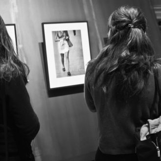 Two people with long hair view a framed photograph on a gallery wall. The black-and-white photo, part of the Magnum collection, captures a person de passage in Paris, walking with a bag in hand.