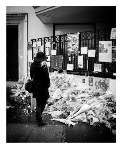 A person dressed in dark clothing stands before a memorial covered with flowers, candles, and messages attached to a fence, paying tribute in a somber, urban setting.