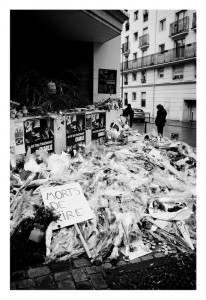A large number of flowers and candles are laid on the pavement as a memorial. Posters line the wall, and a handwritten sign reads "Morts de Rire." Two people stand nearby in the background. The scene is in black and white.