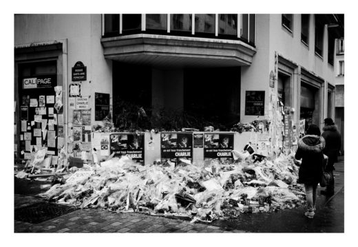 A black-and-white photo shows a street memorial with piles of flowers, candles, and posters reading "Charlie." Notes cover the surrounding walls. Two people walk by on a wet sidewalk, wearing coats.