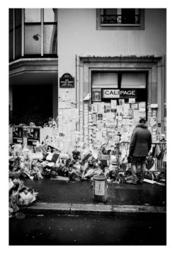 A person stands in front of a building covered with flowers, candles, and notes, likely part of a memorial. The scene is in black and white, and the street appears wet from rain.