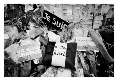 A black and white photo shows a memorial with "Je Suis Charlie" signs, flowers, and handwritten notes placed together in tribute to the Charlie Hebdo attack victims.