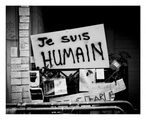 A black and white photo shows a large sign reading "Je suis HUMAIN" amid fences and handwritten notes posted on a wall, conveying a message of solidarity and humanity.