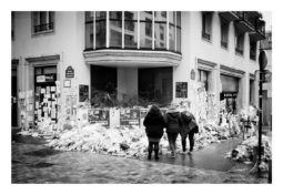 Three people stand on a wet cobblestone street, observing a building covered in posters and surrounded by piles of trash and debris. The scene appears somber and chaotic, with papers and signs visible on the walls.