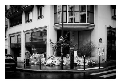 Black and white photo of a street corner with a memorial of flowers and tributes placed on the sidewalk outside a building. A person with an umbrella walks nearby on the rainy street.