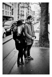 A woman and a man stand on a wet city sidewalk. The woman wears a dark coat and hat, while the man wears a hooded jacket and cap. Parked cars and buildings line the street in the background. The photo is black and white.