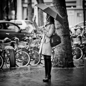 A woman stands on a wet city sidewalk, pluie falling softly as she holds a polka-dotted umbrella and snaps an autoportrait with her phone. Bicycles are parked in the background, and cars line the street in this black-and-white image.