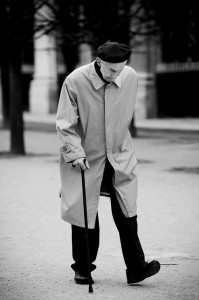 An elderly person in a béret sombre and imperméable clair walks with a cane on a quiet, tree-lined path, head slightly bowed.