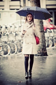 A woman in a beige coat and black tights walks on a wet street, holding a navy blue parapluie à pois blanc. She carries a red bag over her shoulder and looks ahead, with bicycles parked in the background.