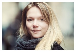 Solène, a young woman with long blonde hair and a soft smile, stands outdoors. She wears a chunky gray scarf and dark jacket, adding a touch of couleur to the blurred background.