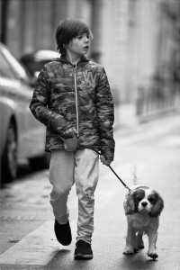 A young boy in a camouflage jacket and sweatpants walks Fred & Rufus, his small dog, on a leash along a city street. The scene is in black and white.