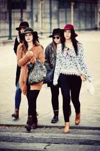 Five young women, known as the gang des chapeaux, stroll confidently down a city street in stylish casual outfits and chic chapeaux en feutre. Their neutral-toned looks stand out against an iron fence and blurred urban background.