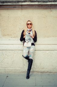Stéphanie Lumion Stéphanie Lumion stands against a light stone wall, wearing sunglasses, a scarf, patterned pants, and black boots, holding a phone and a bag as she looks at the camera.