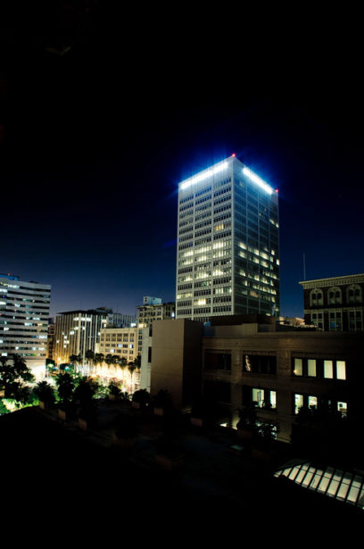A nighttime cityscape featuring a tall, illuminated skyscraper with a glowing peak in Downtown Los Angeles. Surrounding buildings are lit with streetlights, and the scene has a tranquil, serene ambiance. The dark sky contrasts with the artificial lights, enhancing the urban atmosphere.