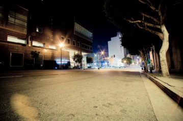 A quiet, empty urban street at night in Downtown Los Angeles is illuminated by streetlights. Buildings line both sides of the street, with some trees near the sidewalk. The late-night scene is tranquil, with a soft glow from the lights reflecting off the pavement.