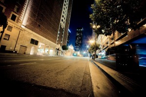 A wide-angle view of a downtown Los Angeles street at night, flanked by tall buildings with illuminated windows. Streetlights and traffic lights brighten the road, while a distant skyscraper glows against the dark sky. Trees line the sidewalks, adding greenery to the late-night urban scene.