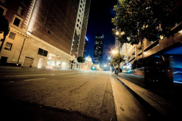 A wide-angle view of a downtown Los Angeles street at night, flanked by tall buildings with illuminated windows. Streetlights and traffic lights brighten the road, while a distant skyscraper glows against the dark sky. Trees line the sidewalks, adding greenery to the late-night urban scene.
