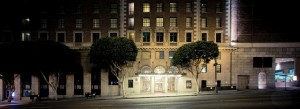 Late night view of an urban building in Downtown Los Angeles with an arched entrance illuminated by bright lights. Large trees flank the entrance. Above, several windows are illuminated, and the street in front is quiet with no visible vehicles or pedestrians.