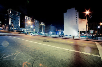 Late-night urban scene featuring an empty street in Downtown Los Angeles with lit buildings in the background. Bright streetlights illuminate the area, casting a soft glow on the surroundings. A large sign is visible on the right side of the image, and a few cars are parked in the distance.
