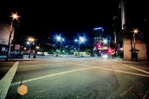 Nighttime city scene featuring an empty intersection, illuminated by streetlights casting a starburst effect. Surrounding buildings are visible, including one with a vertical sign on the right. Traffic signals show red, with a clear, dark sky above in late-night Downtown Los Angeles.