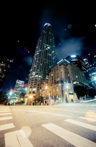 A nighttime cityscape in Downtown Los Angeles features tall, illuminated skyscrapers and a brightly lit intersection with blurred traffic lights, capturing the dynamic energy of the urban environment. The sky adds a dramatic backdrop with a mix of clouds and light effects, epitomizing late-night vibrancy.