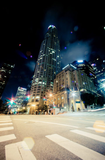 A nighttime cityscape in Downtown Los Angeles features tall, illuminated skyscrapers and a brightly lit intersection with blurred traffic lights, capturing the dynamic energy of the urban environment. The sky adds a dramatic backdrop with a mix of clouds and light effects, epitomizing late-night vibrancy.