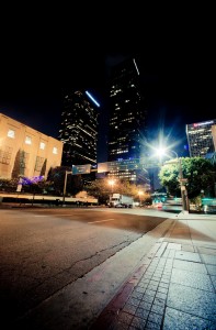 A nighttime cityscape of Downtown Los Angeles features tall, illuminated skyscrapers with lighted windows. The street in the foreground, bathed in light from streetlights, sees a few cars and a bus driving by. Trees and building facades line the road, enhancing the late-night urban atmosphere.