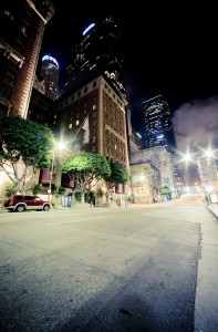 A nighttime city street view in Downtown Los Angeles features tall buildings illuminated by lights. Streetlights cast a glow over the road with a few cars parked along the side. Trees line the sidewalk, and the buildings display a mix of architectural styles against a dark sky, capturing the late-night vibe.
