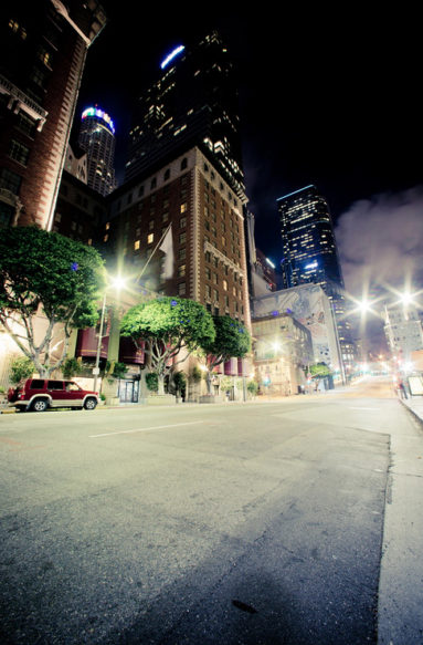 A nighttime city street view in Downtown Los Angeles features tall buildings illuminated by lights. Streetlights cast a glow over the road with a few cars parked along the side. Trees line the sidewalk, and the buildings display a mix of architectural styles against a dark sky, capturing the late-night vibe.
