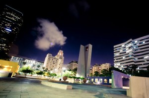 Late-night cityscape of Downtown Los Angeles featuring modern skyscrapers and distinctive architecture, including a tall clock tower and buildings with illuminated windows. The plaza includes large spherical structures and benches, with a partly cloudy night sky overhead.