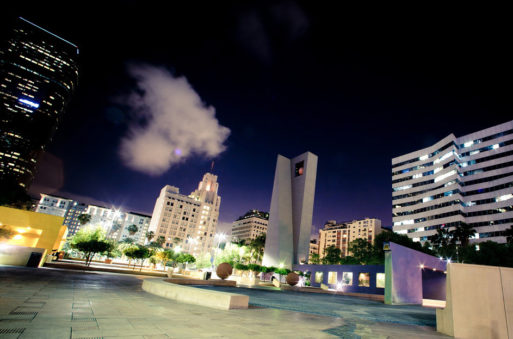 Late-night cityscape of Downtown Los Angeles featuring modern skyscrapers and distinctive architecture, including a tall clock tower and buildings with illuminated windows. The plaza includes large spherical structures and benches, with a partly cloudy night sky overhead.