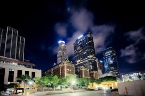 A cityscape of downtown Los Angeles at late night, featuring illuminated skyscrapers and a well-lit foreground with trees. The sky above is dotted with clouds, and lights from the buildings create a vibrant urban atmosphere.