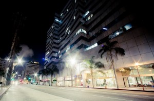 A city street in Downtown Los Angeles at night with tall, illuminated office buildings. The bright streetlights and building lights reflect off the structures. Palm trees line the street, and the late-night scene has an empty, quiet atmosphere.