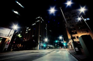 A deserted downtown Los Angeles street at night is illuminated by streetlights, casting a bright glow on the surroundings. Tall buildings on both sides showcase numerous lit windows, and a zebra crossing is visible in the foreground. The late-night scene is calm and quiet.