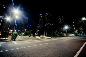 Wide-angle night view of a deserted city street in Downtown Los Angeles illuminated by bright streetlights. Tall buildings with lit-up windows line the street, with a few palm trees on the right. The empty road and the cityscape create a serene and calm atmosphere on this late night.