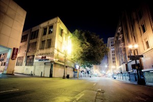 A nighttime view of an empty urban street with old buildings, some with lit signs. A large tree is in front of a building on the left, and a "Parking" sign is visible. The street is well-lit, and the scene has a calm, serene ambiance indicative of late-night Downtown Los Angeles.