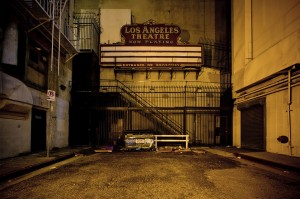 A dimly lit alley in downtown Los Angeles with a vintage sign reading "Los Angeles Theater - Now Playing." The entrance is gated and the marquee is empty, casting an eerie glow late at night. The alley is flanked by tall buildings and cluttered with debris, including a wooden pallet and a dumpster.