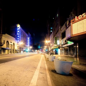 A brightly lit, empty city street at night with illuminated signs, including "Palace" and "Los," on both sides. Several large planters line the sidewalk, and the buildings display classic architectural details of Downtown Los Angeles, with neon lights reflecting off the pavement in this late-night scene.