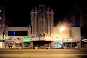 A historic theater with ornate architecture stands closed at night, its marquee unlit. The surrounding storefronts, including a boutique and restaurant, are dimly illuminated by streetlights in downtown Los Angeles. The late-night scene exudes a nostalgic, almost eerie ambiance.