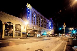 Nighttime view of a street in downtown Los Angeles with well-lit buildings. A large building with prominent signage reading "PALACE" is highlighted, adorned with bright blue lights. Other smaller buildings and streetlights illuminate the empty late-night street, where some signage and advertisements are visible.