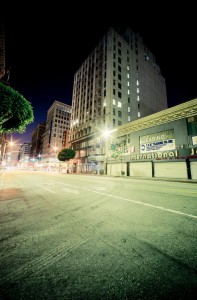 A late-night view of an empty city street in downtown Los Angeles, flanked by towering buildings. The illuminated structures sport signs and billboards, including one for a leasing office. Streetlights cast a greenish glow, and the scene is devoid of people or traffic.