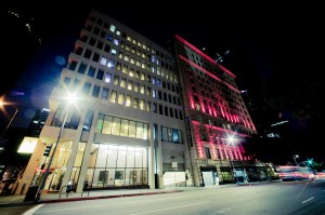 A nighttime cityscape in Downtown Los Angeles features a street with two prominent buildings. The left building boasts a modern design with a lit-up interior, while the right building has a classic façade illuminated with vibrant red lights. Cars are seen driving past in the late night ambiance.