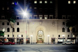 Street view of a large, ornate building entrance with an arched, illuminated doorway flanked by columns in downtown Los Angeles. The facade has multiple windows and palm trees on each side. Cars are parked along the street, and a few street lamps are alight in the late night.