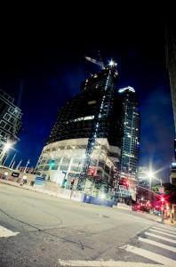 A high-rise building under construction at night, illuminated by construction lights. A crane is visible atop the structure. Surrounding buildings and streetlights enhance the urban nightscape of Downtown Los Angeles, with traffic lights emitting a red glow at an empty intersection in the late-night foreground.
