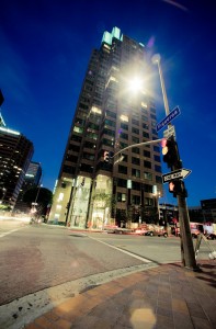 A nighttime view of a tall, illuminated skyscraper at a Downtown Los Angeles intersection. Street signs read "Figueroa" and "Do Not Enter." Surrounding buildings are partially visible, with streetlights and light trails from passing cars enhancing the late night urban scene.