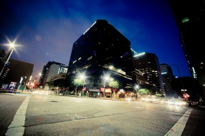A late-night cityscape of Downtown Los Angeles captures a deserted urban intersection illuminated by streetlights. Tall buildings with lit windows surround the area, and the sky is deep blue transitioning to cloudy purple. Red, green, and blue street markings are visible on the pavement.