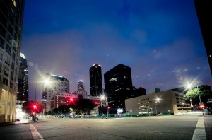 A city street at night with tall buildings illuminated by streetlights and city lights. The sky is a deep blue, and the street is empty, giving a serene and peaceful atmosphere. Several high-rise buildings form the urban skyline of Downtown Los Angeles in the background on this late night.