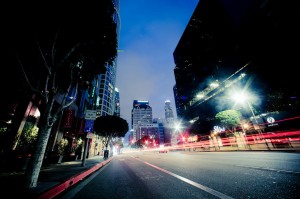 A city street at twilight with skyscrapers illuminated in the background offers a mesmerizing view of Downtown Los Angeles. Light trails from passing cars create streaks of red and white. Trees line the sidewalks, and a mix of illuminated and dark buildings frame the scene beautifully.
