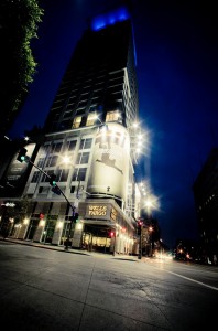 In Downtown Los Angeles, a city street at night showcases a tall, illuminated building with a Wells Fargo sign and a large advertisement of a silhouetted athlete in a dynamic pose. The street is empty, bathed in the glowing light of traffic lights, capturing the tranquility of the late night scene.
