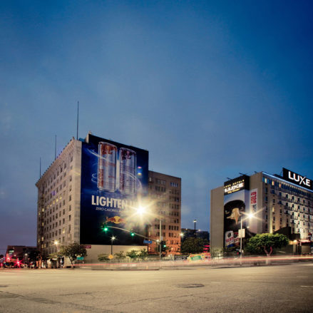A cityscape at dusk in Downtown Los Angeles features illuminated billboards on buildings. One building displays an advertisement for a beverage with the text "LIGHTEN UP." The streets are relatively empty, and lights from buildings and cars create a serene evening atmosphere.